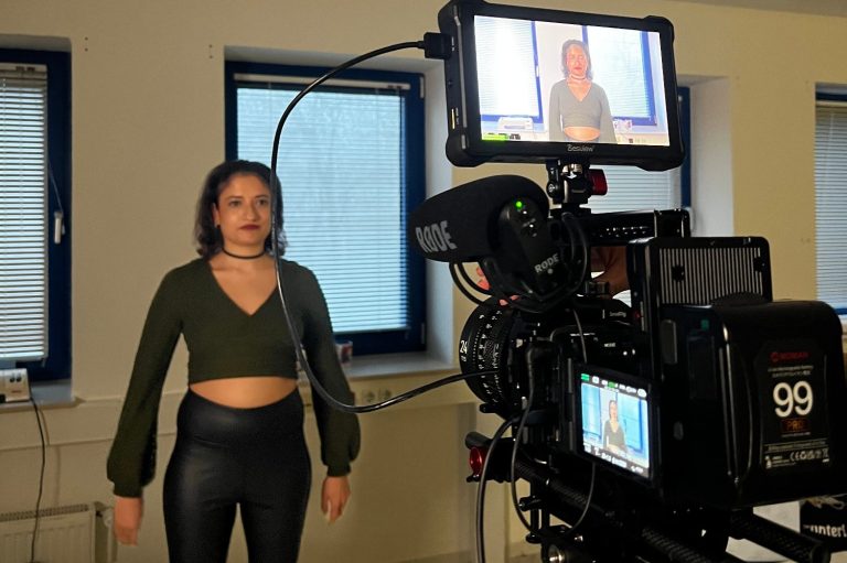 Frau in grünem Oberteil und engen, schwarzen Hosen vor einer Kamera bei einer Filmproduktion
